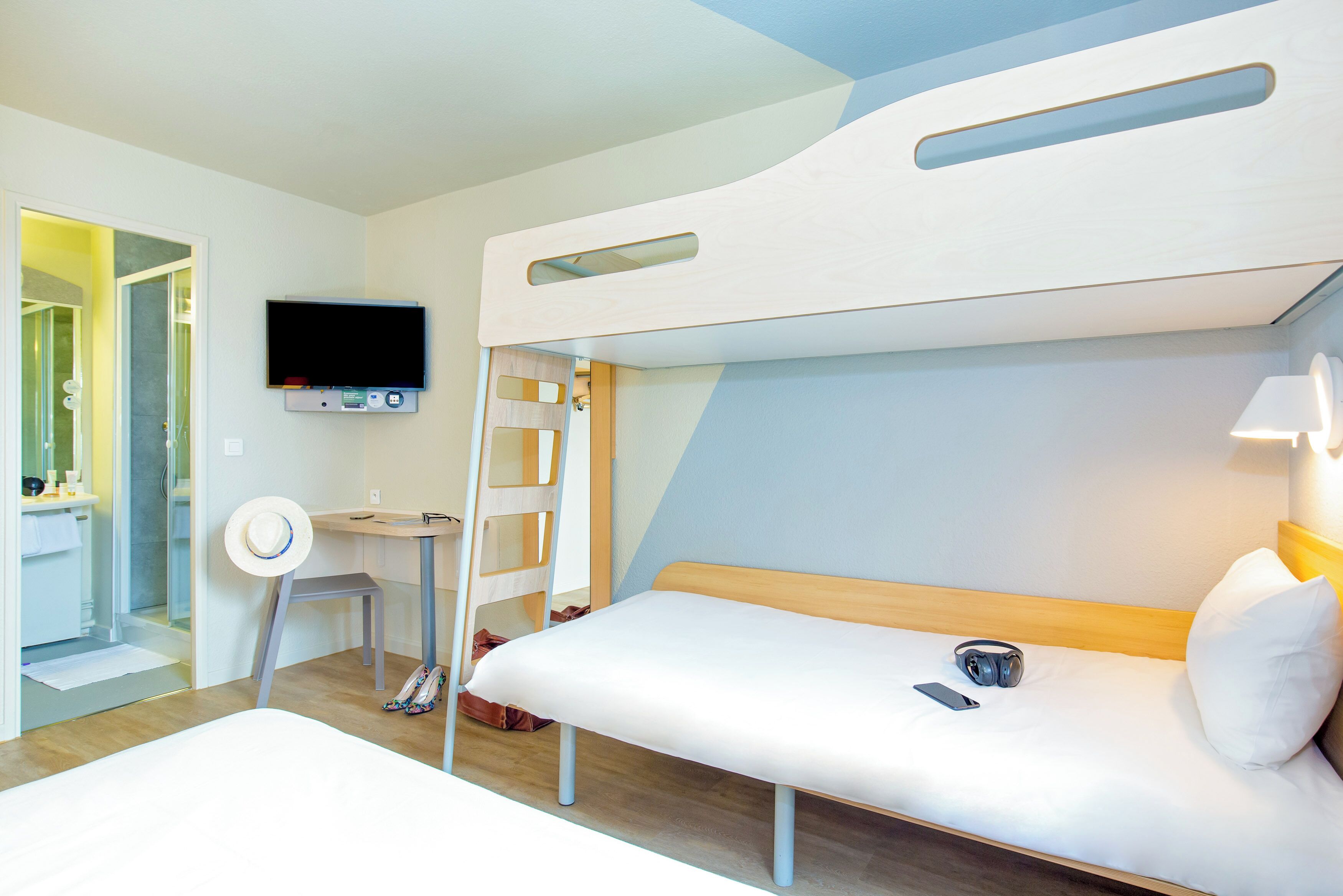 Quarto de hotel moderno com beliche, escrivaninha, cadeira, televisão e banheiro visível por uma porta aberta.