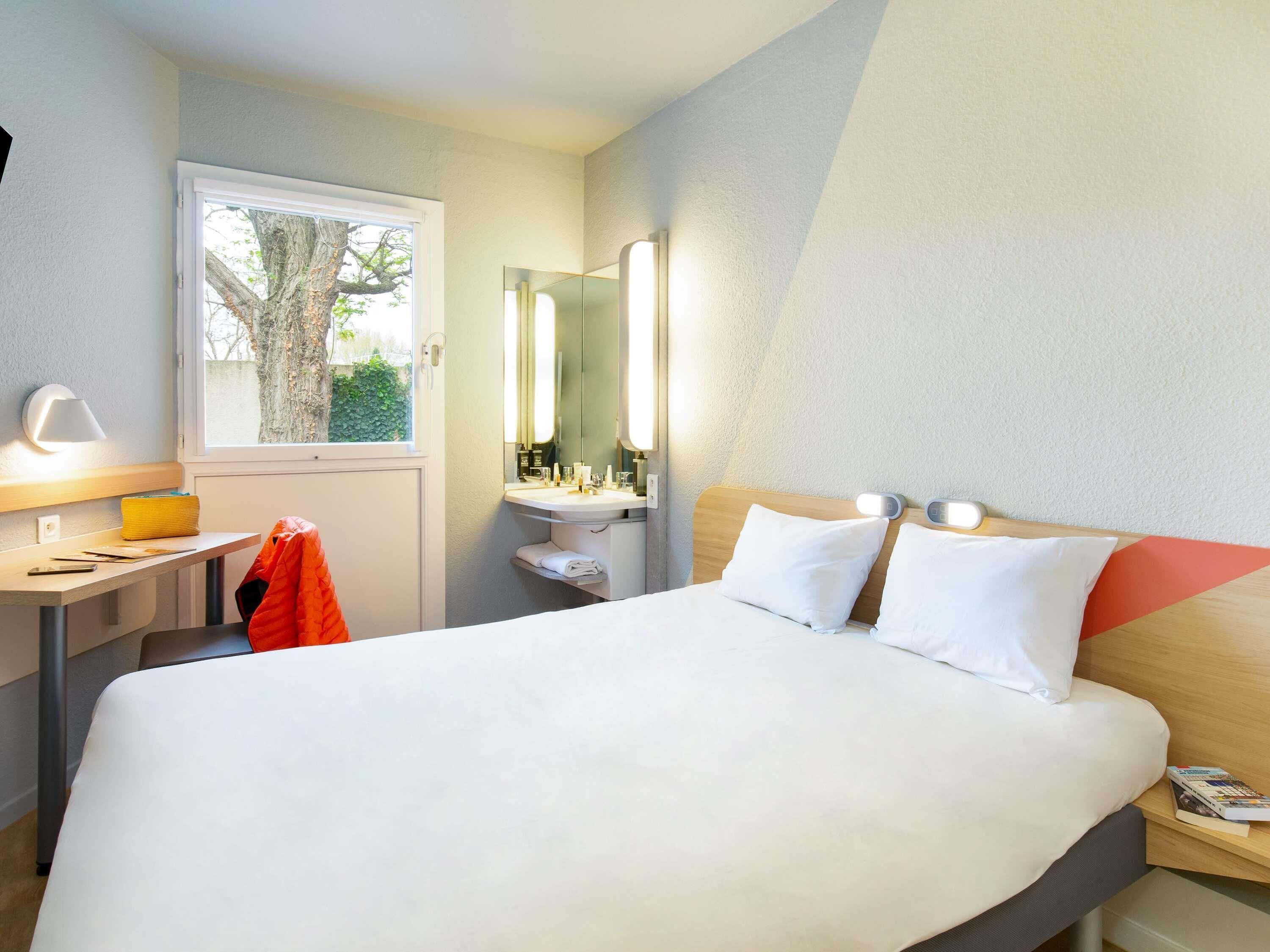 A hotel room with a bed, bedside table, lamp, mirror, and window.