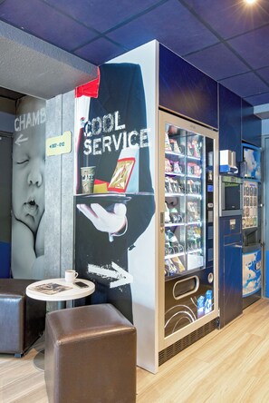 Un interior moderno con una máquina expendedora que dice 'Cool Service', una zona de estar con una mesa y una pared con un anuncio grande.