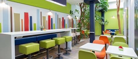 Een modern café met een bar, zitplaatsen in groen en oranje, en een kleurrijk wandontwerp.