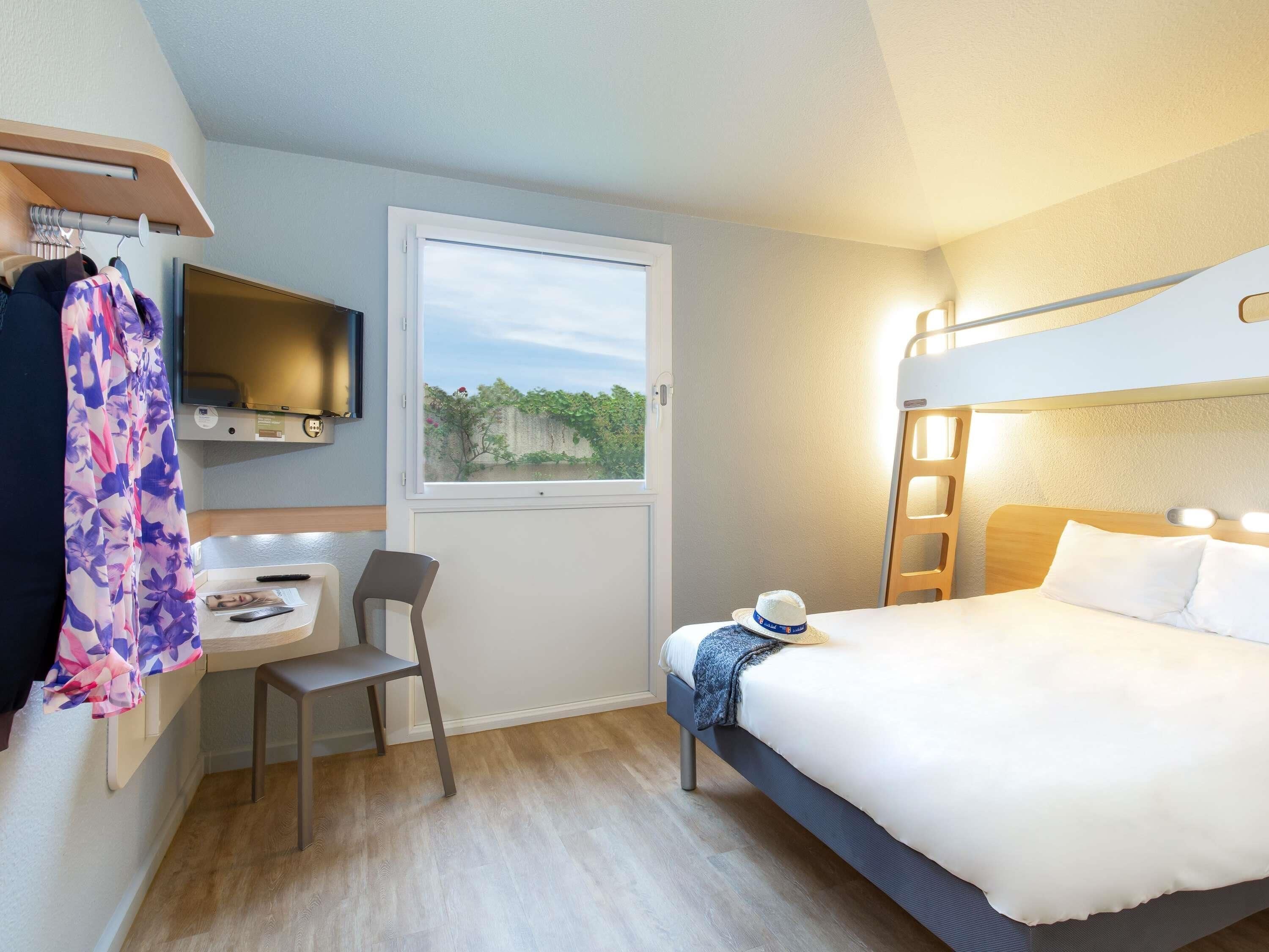 A compact hotel room with a bunk bed, a desk with a chair, a television mounted on the wall, and a window with a view of greenery.
