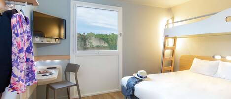 A compact hotel room with a bunk bed, a desk with a chair, a television mounted on the wall, and a window with a view of greenery.