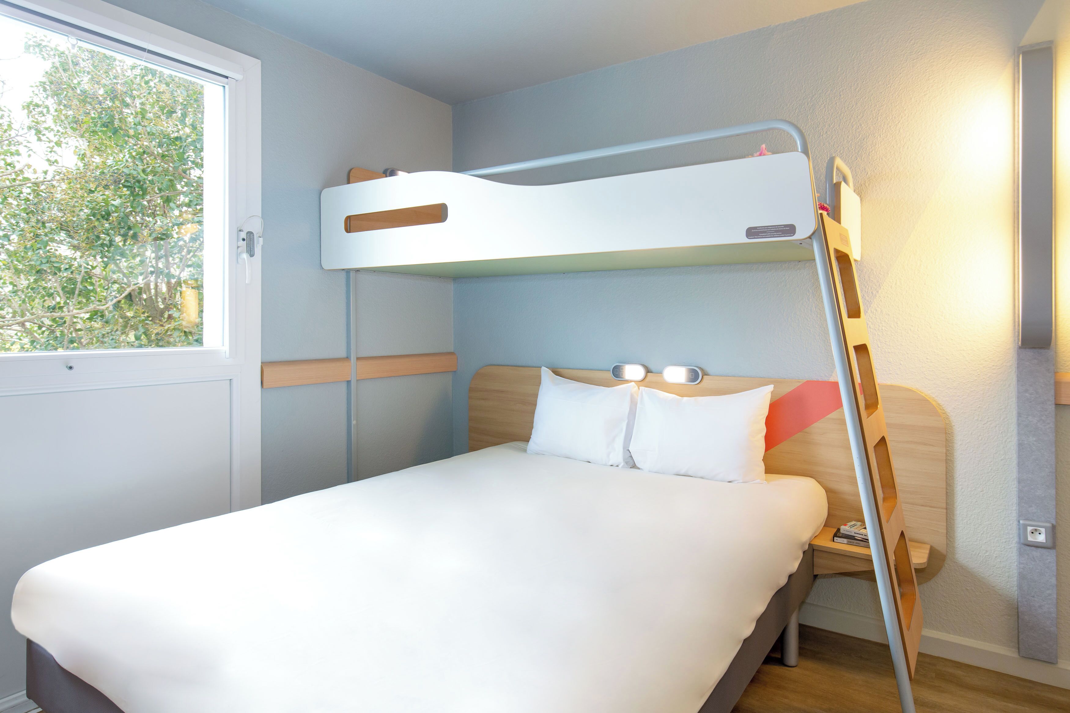 A bunk bed with a ladder, a bedside table, and a window with a view of trees.