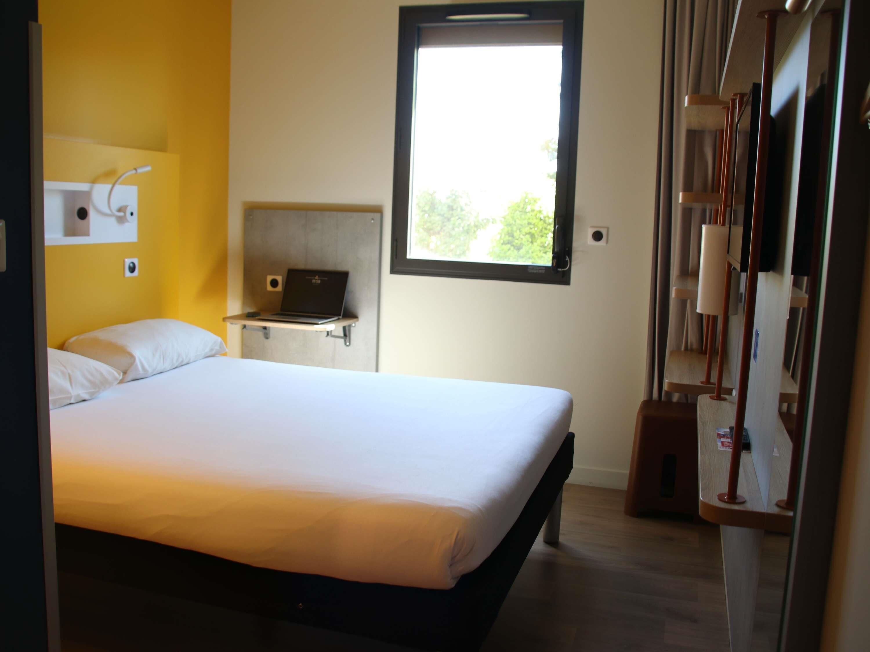 A bedroom with a bed, a laptop on a shelf, a window, and a wardrobe.