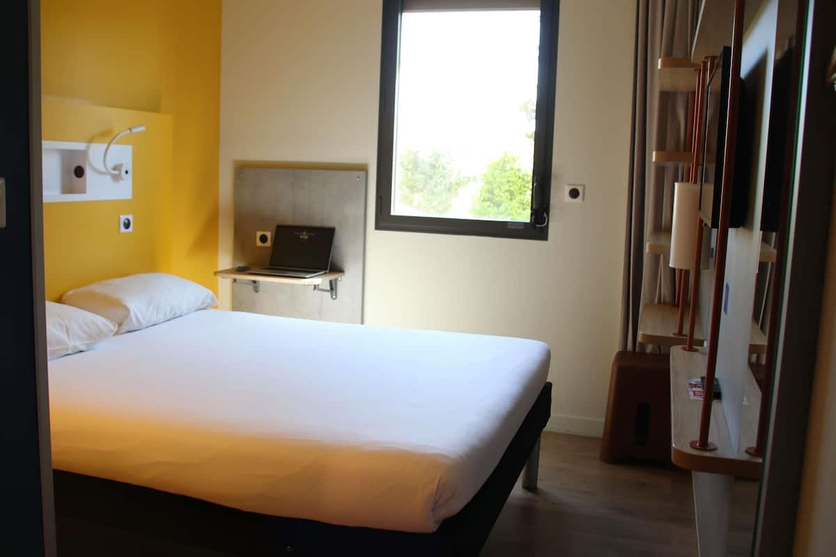 Ein Schlafzimmer mit einem Bett, einem Laptop auf einem Regal, einem Fenster und einem Kleiderschrank.