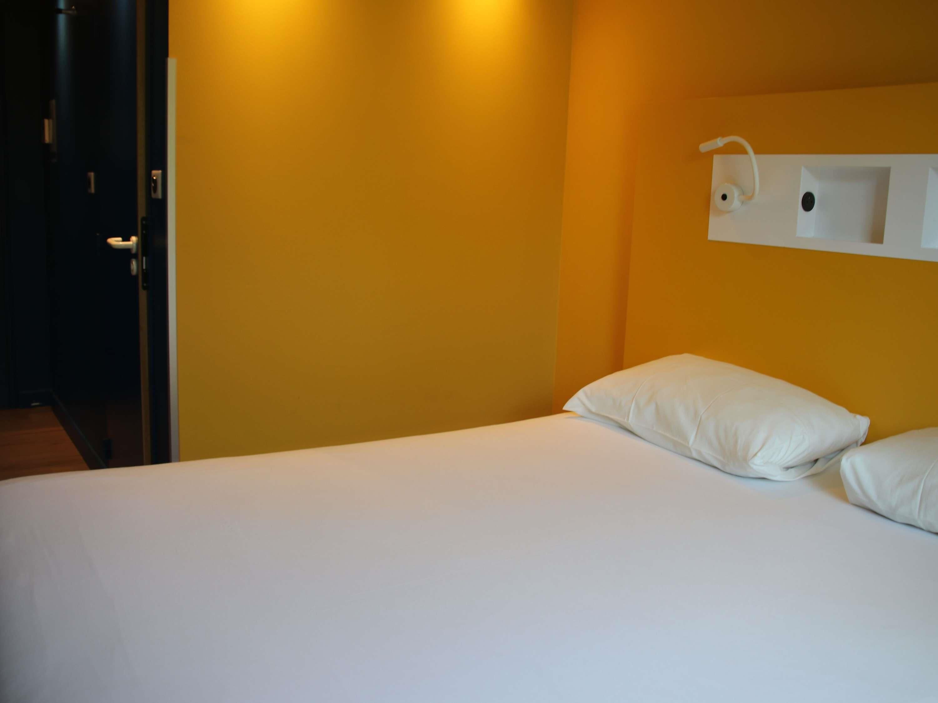 Ein Hotelzimmer mit einem Bett, weißen Kissen, einem Nachttisch mit einer Lampe und einer Tür links.
