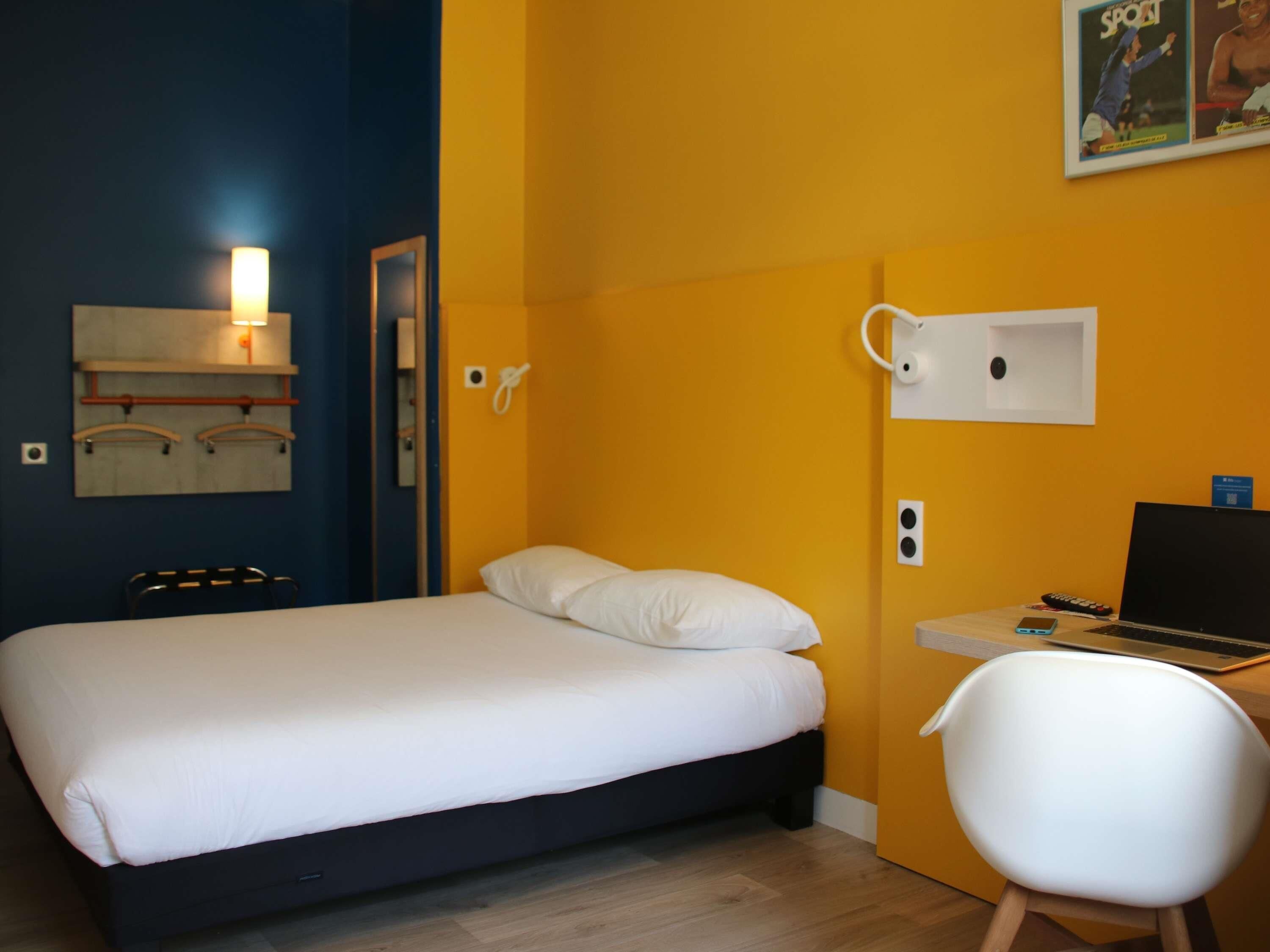 A bedroom with a bed, a desk with a laptop, a chair, and a wall-mounted phone.