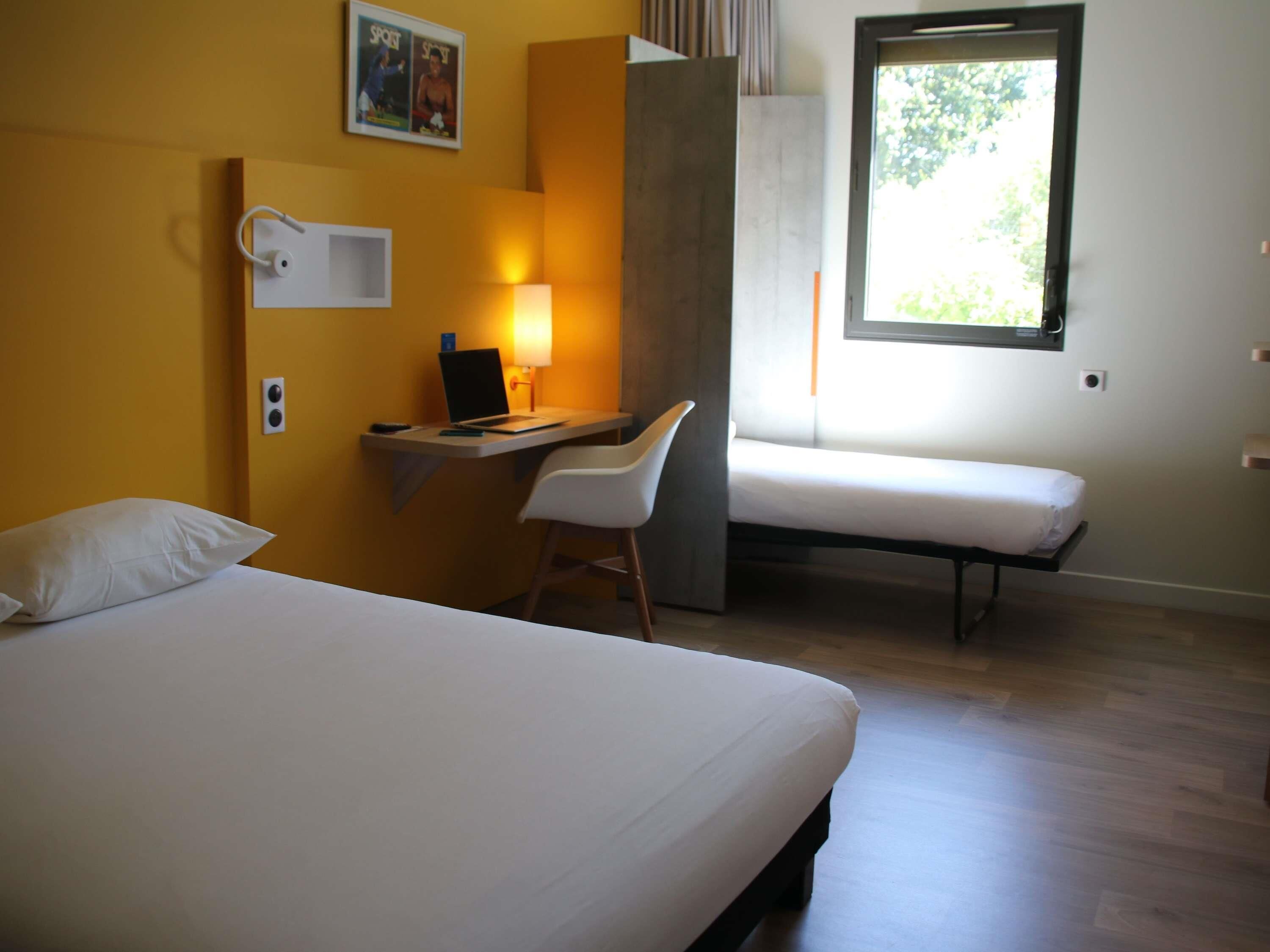 A hotel room with a bed, a desk with a chair, a window, and a wall-mounted light.