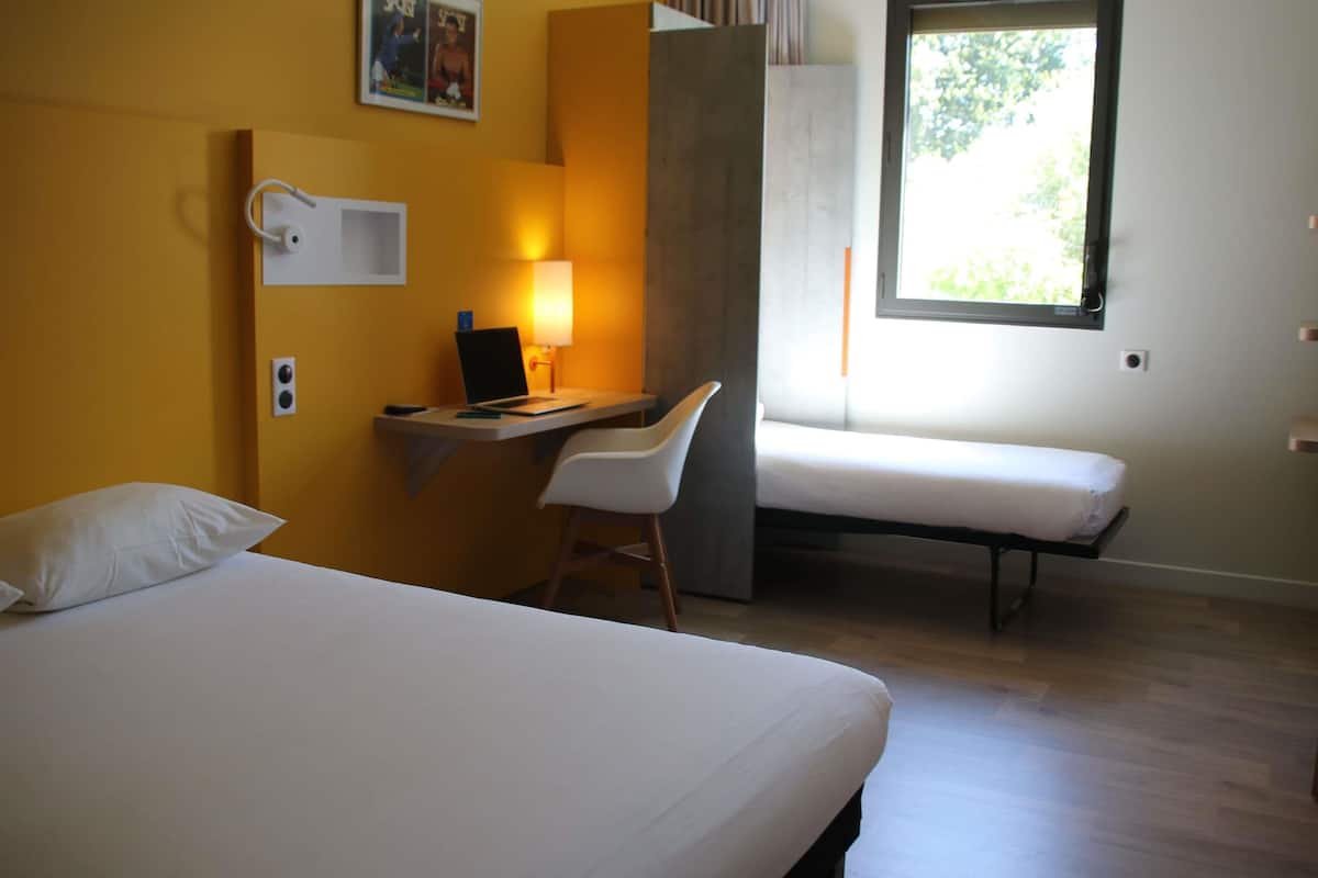 Ein Hotelzimmer mit einem Bett, einem Schreibtisch mit Stuhl, einem Fenster und einer Wandlampe.