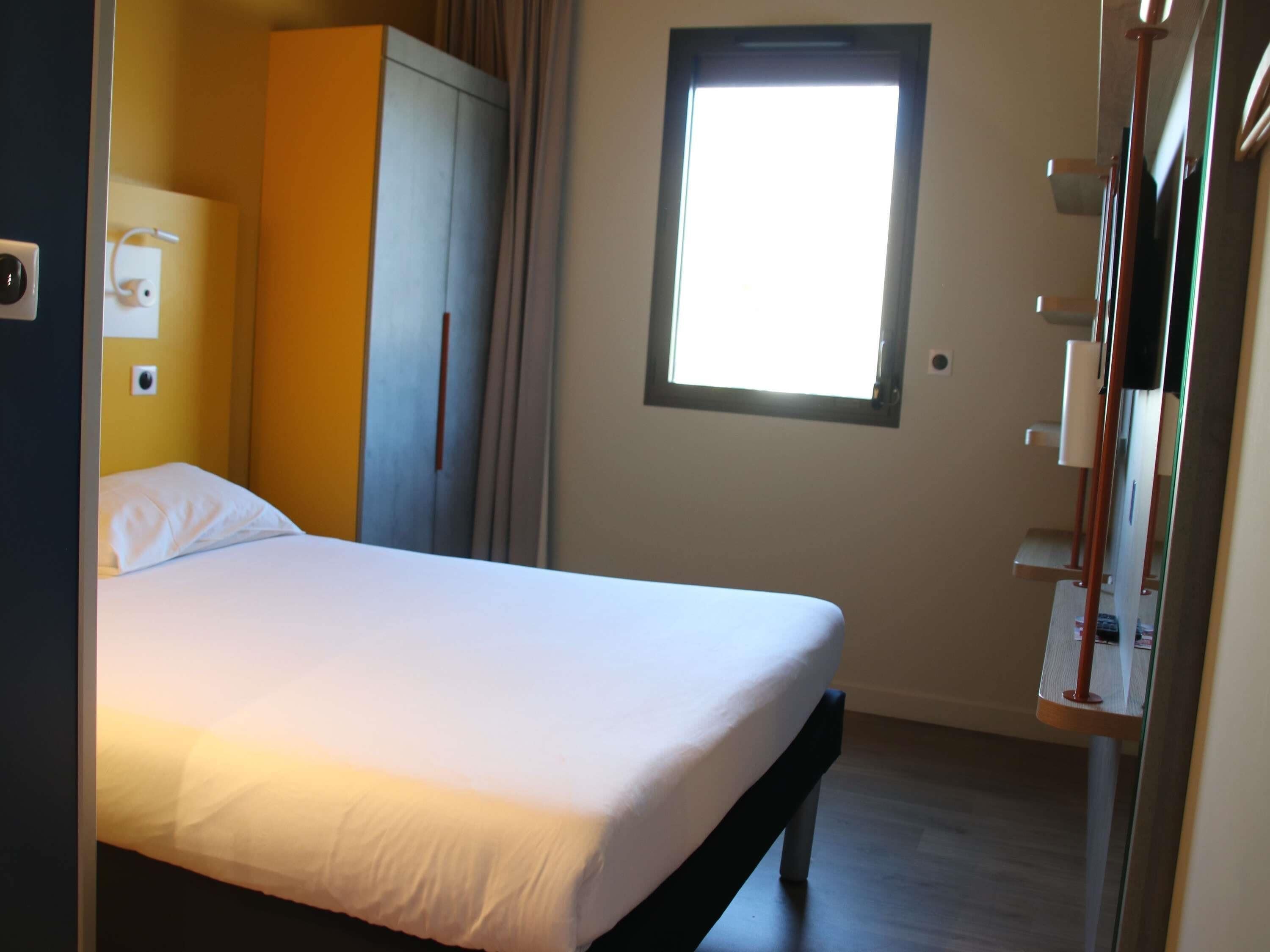 Una cama individual con colchón blanco y cabecero de madera, un armario, una ventana con luz natural y un estante con espejo.