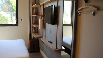 A hotel room with a bed, a TV mounted on the wall, a small stool, and a window with curtains.