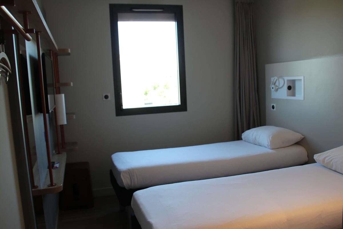 Une chambre d’hôtel avec deux lits, une fenêtre, un miroir et un interrupteur fixé au mur.