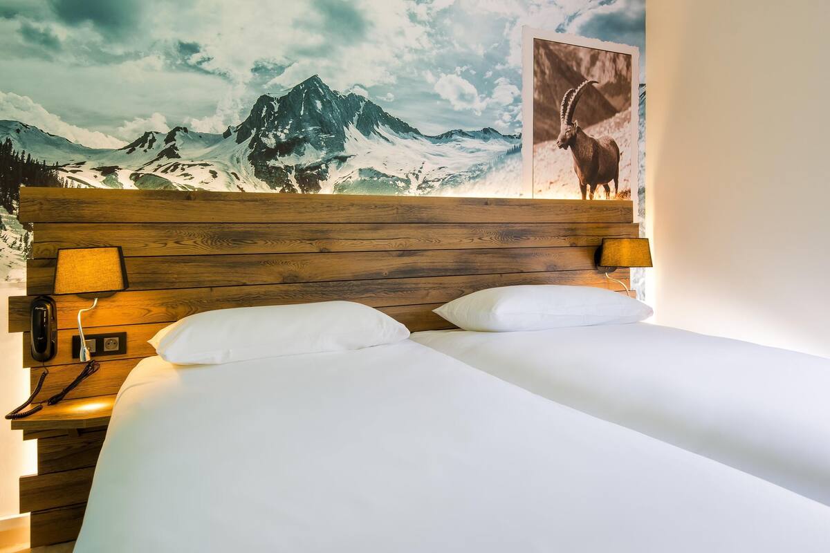 A hotel room with a wooden headboard, two white pillows, a bedside lamp, a telephone, and a mountain view mural.