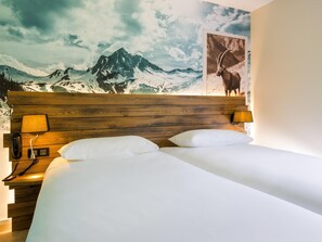 Une chambre d’hôtel avec une tête de lit en bois, deux oreillers blancs, une lampe de chevet, un téléphone et une fresque murale représentant un paysage de montagne.