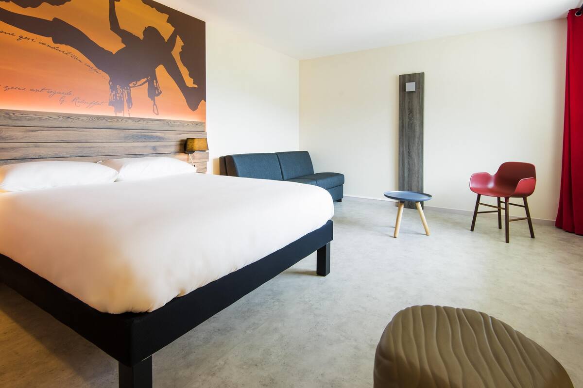 A hotel room with a bed, a blue sofa, a red chair, a small table, and a wall art piece.