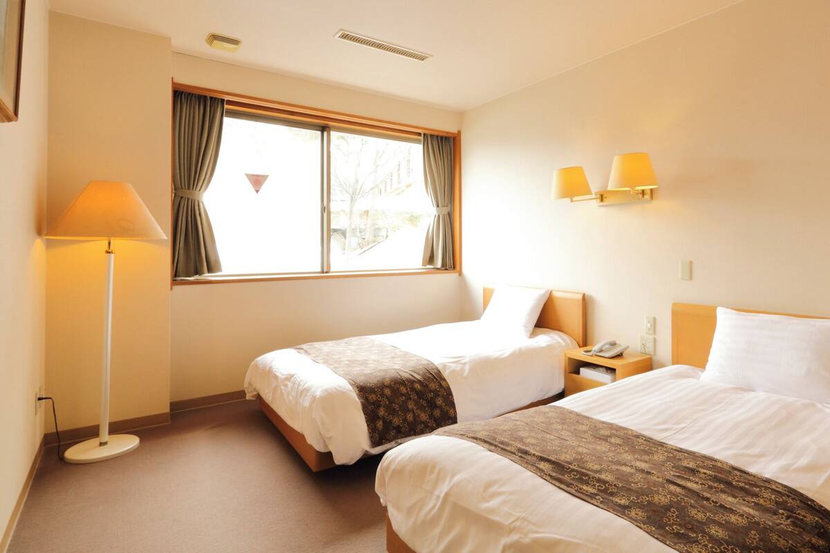 Habitación de hotel con dos camas, una mesita de noche con lámpara, una ventana con cortinas y una lámpara de pared.