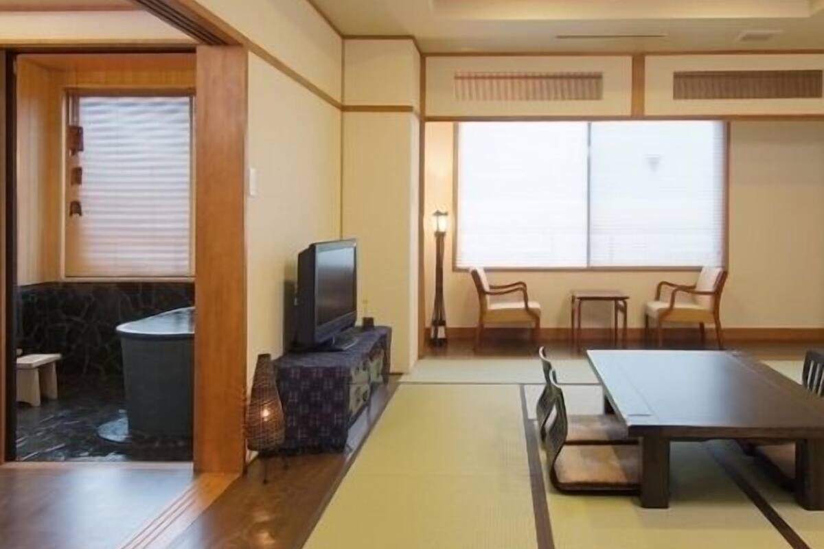 Ein traditioneller japanischer Raum mit Tatami-Boden, einem niedrigen Tisch, Stühlen, einem Fernseher und einem Fenster mit Jalousien.