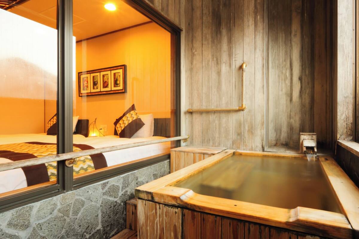 Ein traditionelles japanisches Onsen mit Holzbad, Steinwänden und einem Bett mit gemusterten Betttextilien.