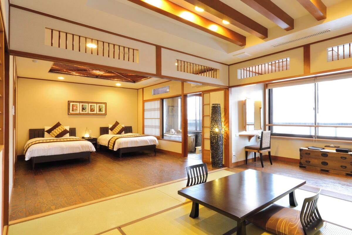Habitación de hotel con dos camas, una mesa de comedor, sillas, un ventanal grande y una lámpara de estilo japonés tradicional.