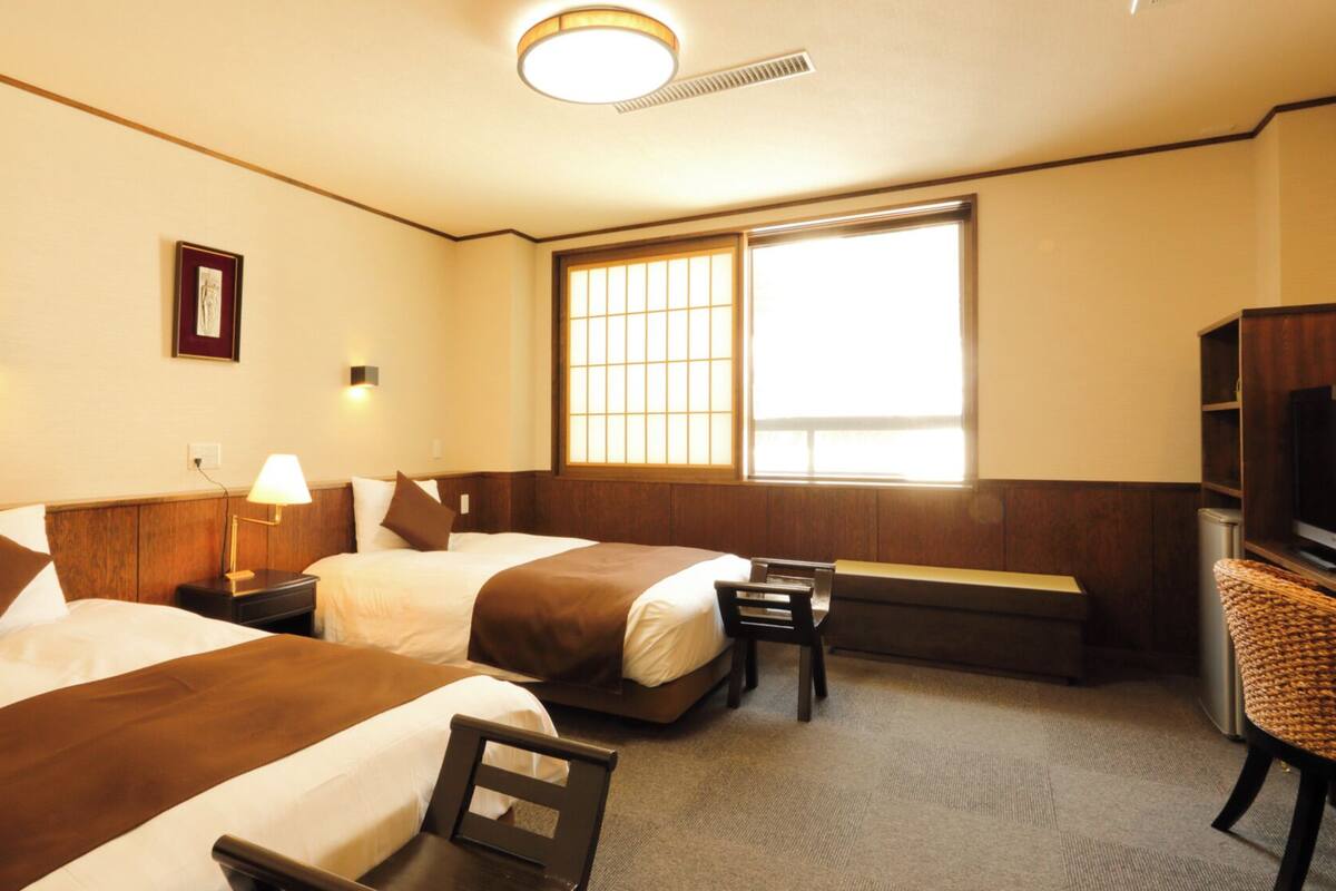 Una habitación de hotel con dos camas, televisión, escritorio y ventana con persianas.