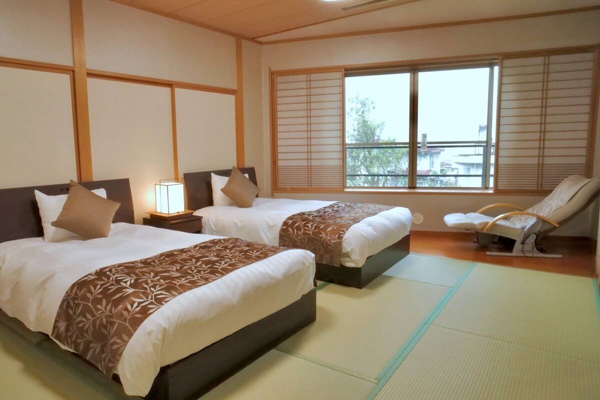 Ein Hotelzimmer mit zwei Betten, einer Sitzecke mit einem Sessel und einem großen Fenster mit Jalousien.