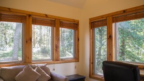 A room with a beige sofa, a black chair, and a small black ottoman. There are three large windows with wooden frames, and the room has a tiled floor. - Riverfront - main level master bedroom - now with high speed unlimited Internet! (Bowden)