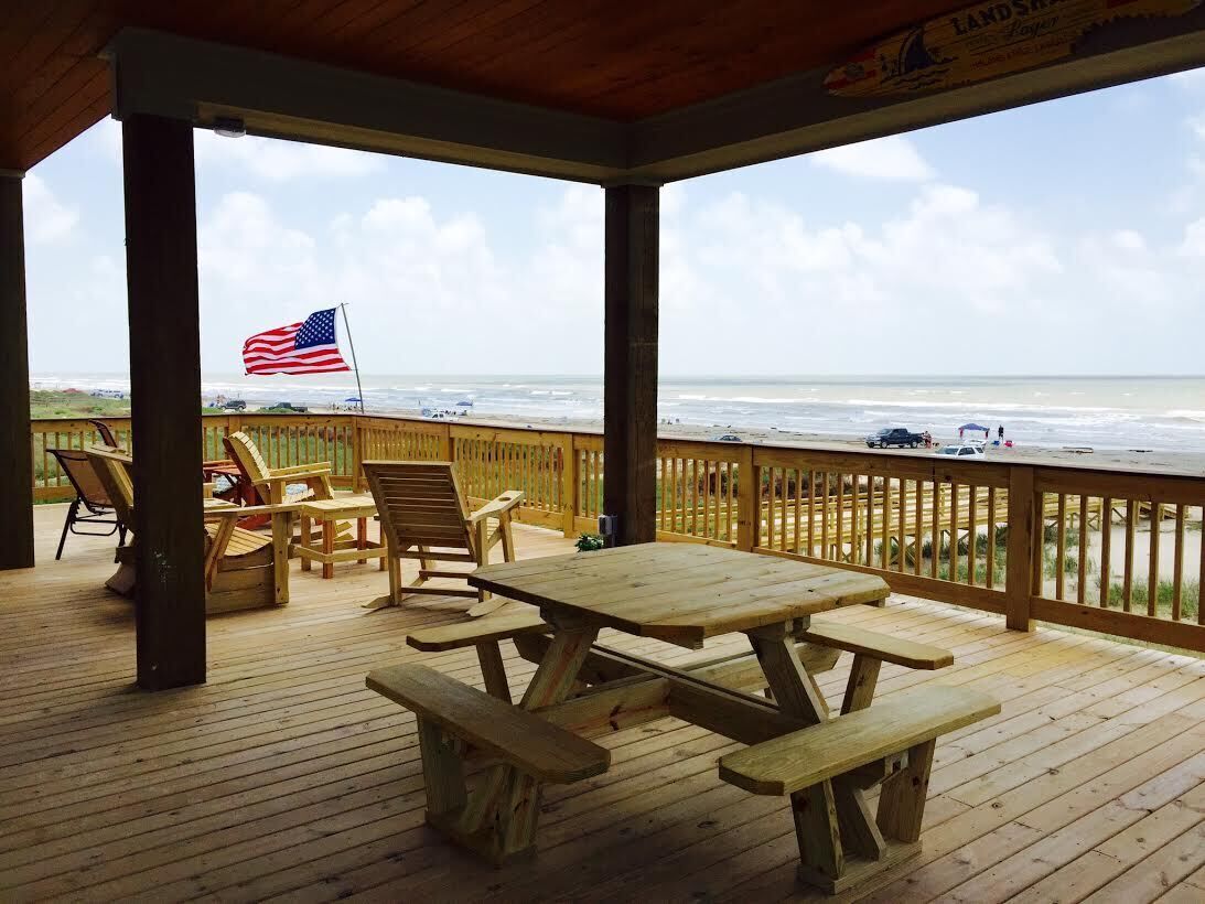 Una terraza de madera con una mesa y sillas de picnic, con vista a una playa y una bandera estadounidense.