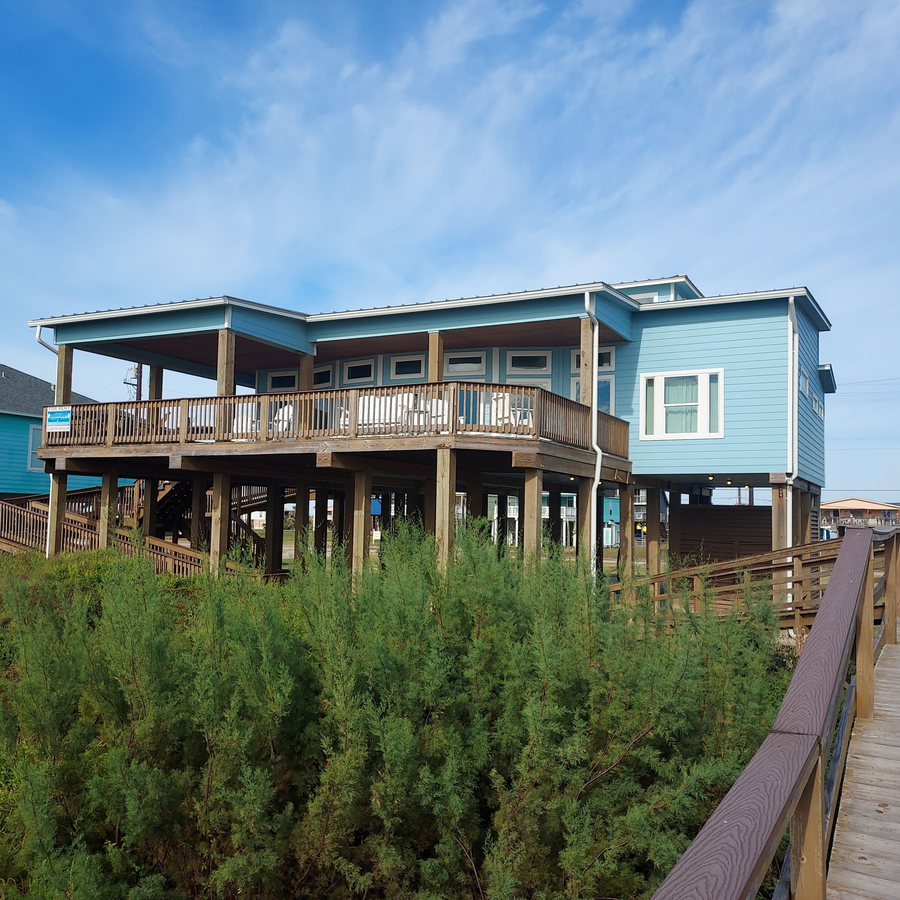 A two-story beachfront house with a wooden deck and a covered porch.