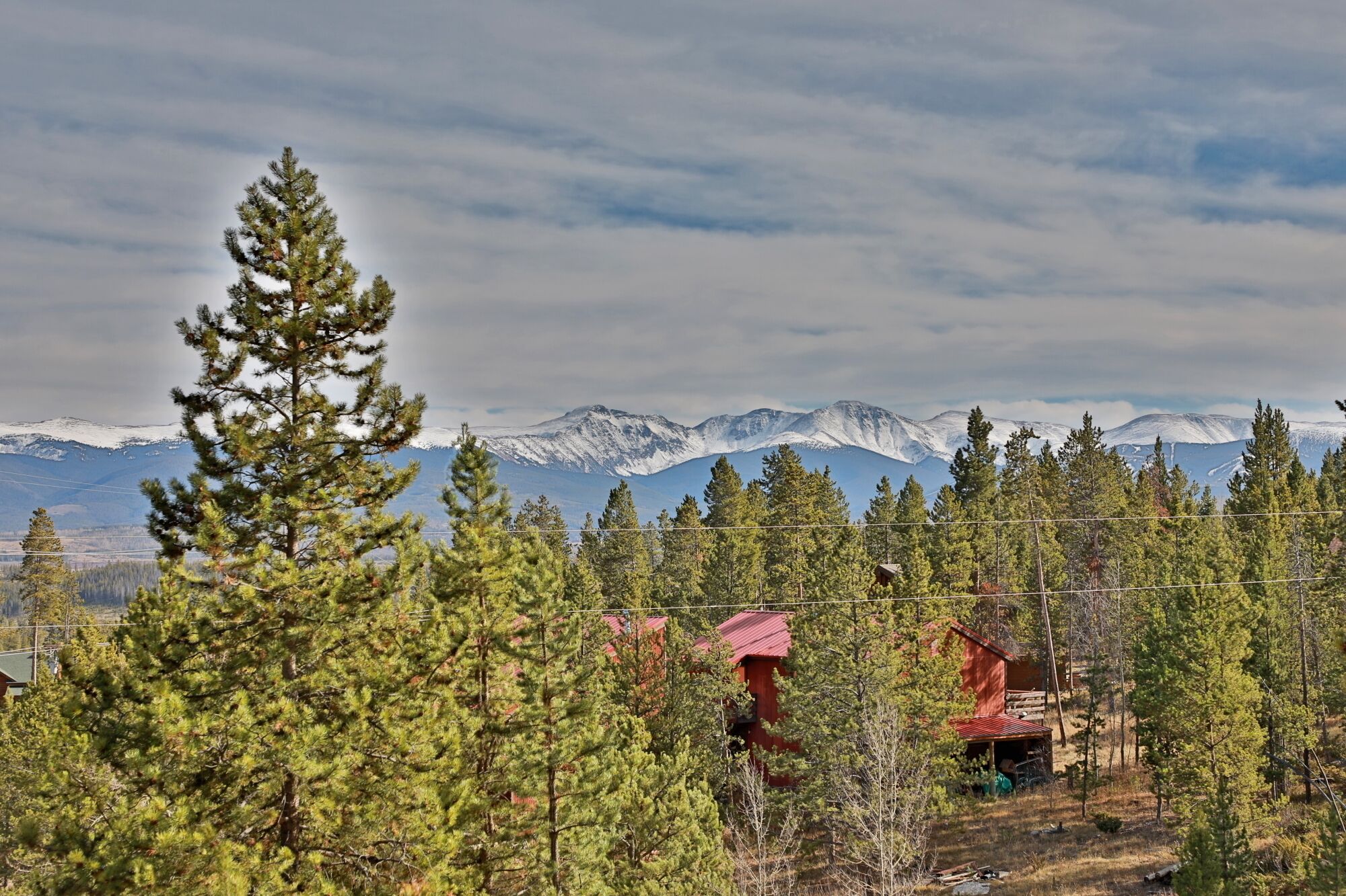 A cabin with a red roof surrounded by dense coniferous forest, overlooking snow-capped mountains.