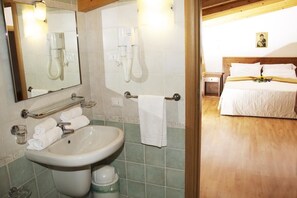 A bathroom with a sink, mirror, and towel rack. There is a bed with white bedding in the adjacent room.