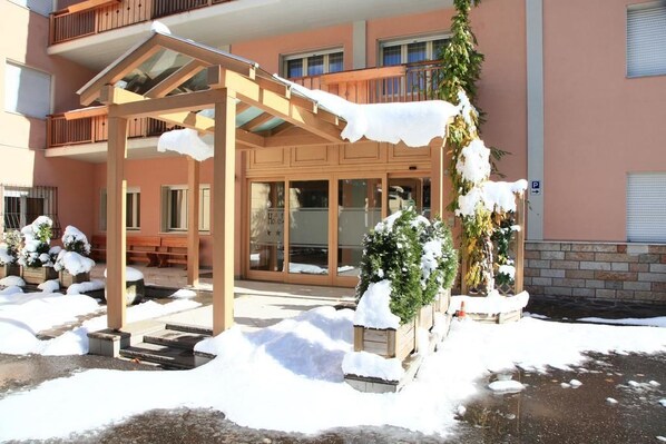 A covered outdoor area with a wooden pergola, snow-covered plants, and a building entrance with glass doors.