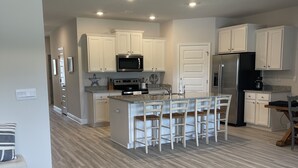 A modern kitchen with white cabinets, a central island with bar stools, stainless steel appliances, and a wooden floor. - The Heron Beach House in Windmark / Mexico Beach, 2 Pools & 5 min walk to Beach. (Port St Joe)
