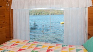 A room with a bed, a window with blinds, and a view of a body of water with boats. - Lucky Catch: Best Cottage on the Best Lake! (Penn Yan)