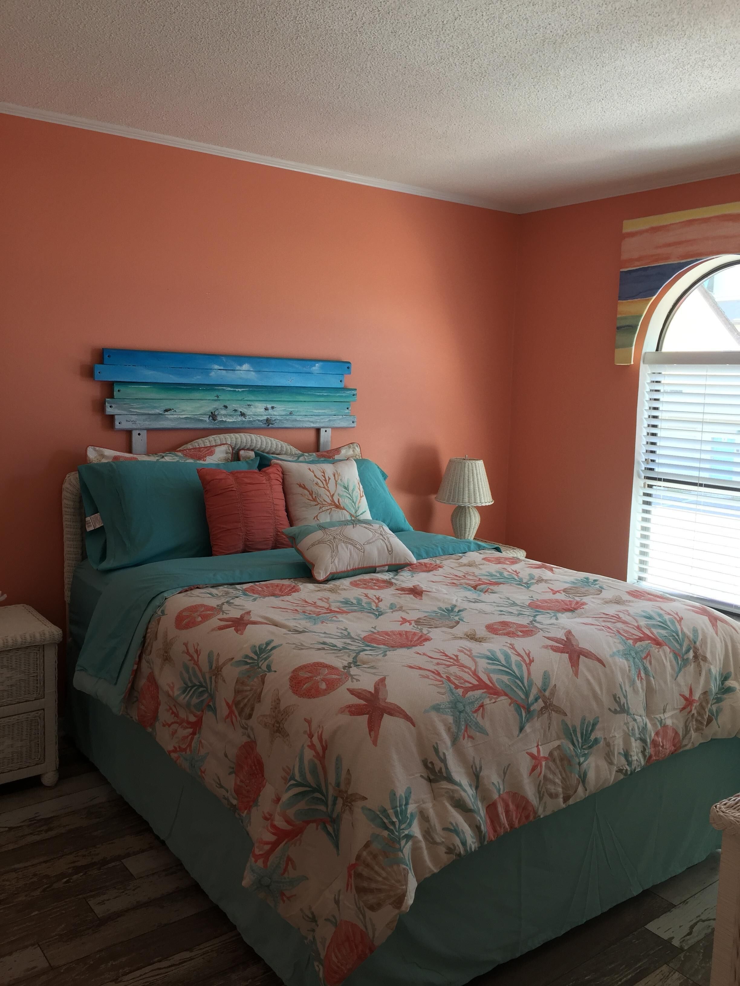 Un dormitorio con cama, cabecera con escena marina pintada, mesita de noche con lámpara y ventana con persianas.