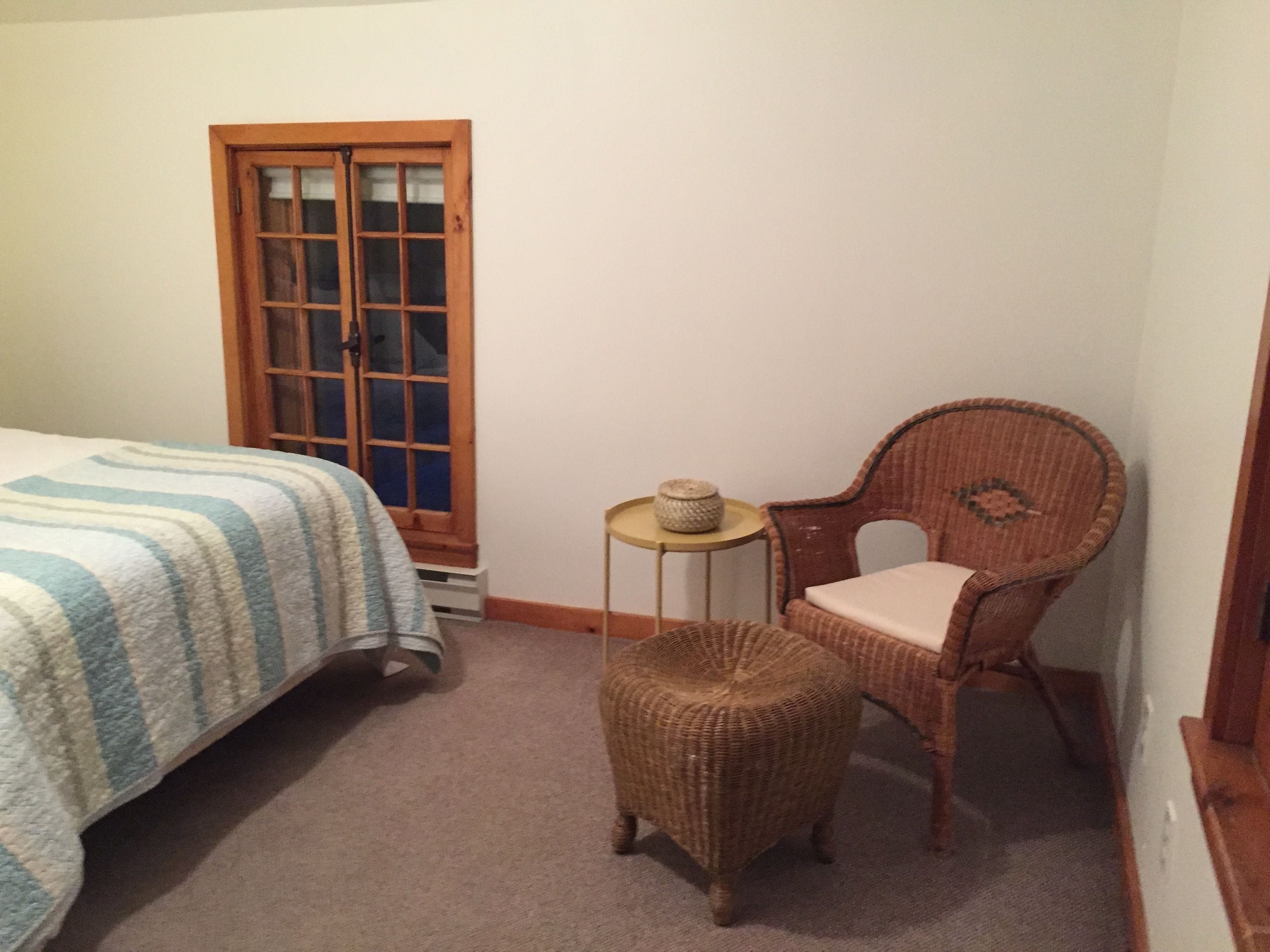 Un dormitorio con una cama, una silla de mimbre, una mesita y un ventanal.