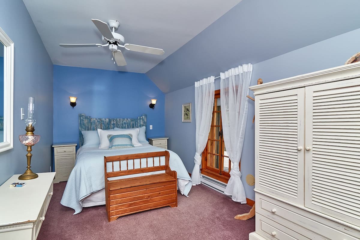 Une chambre à coucher comprenant un lit, une table de chevet, une commode, un ventilateur de plafond et une fenêtre avec des rideaux.