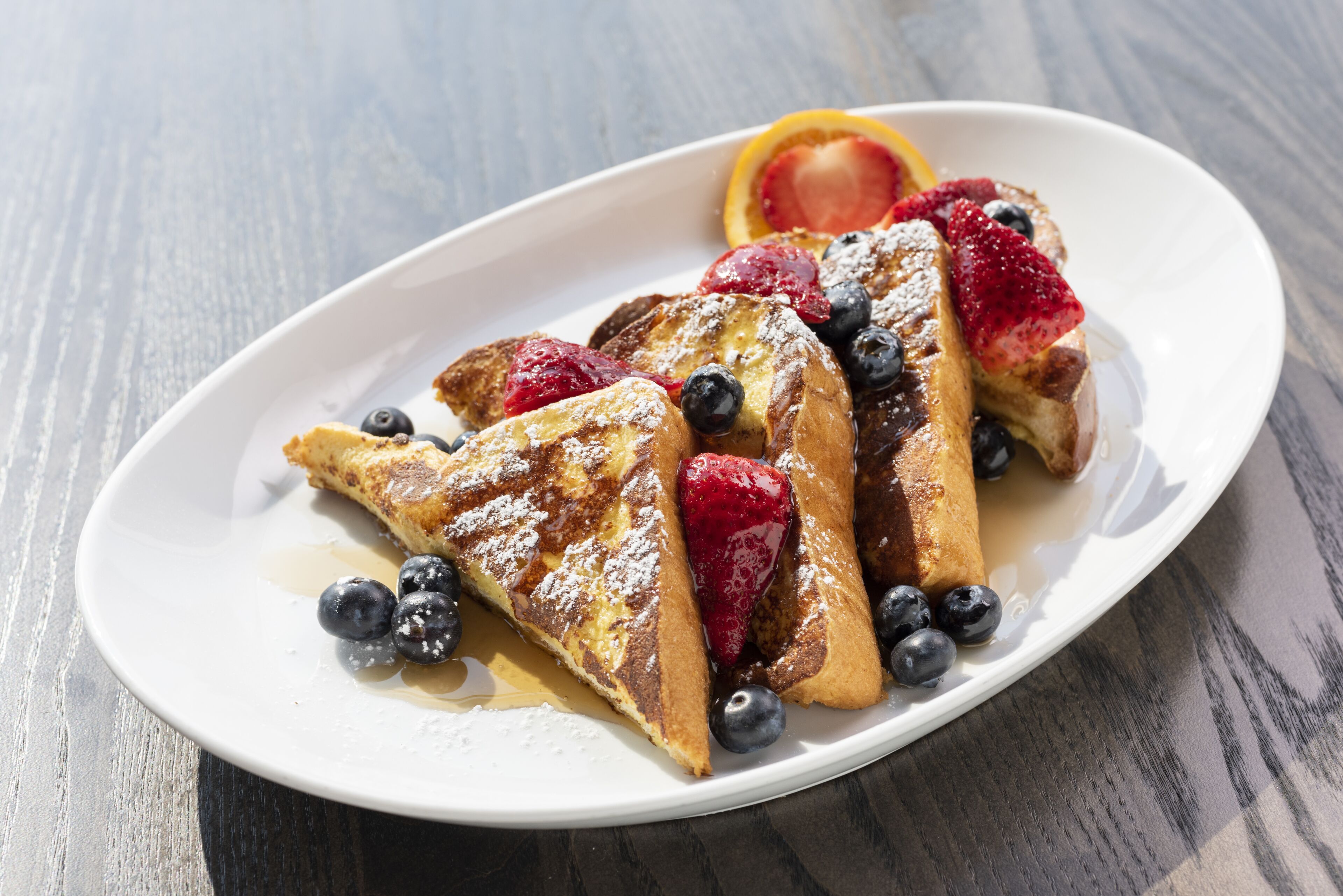 A white plate with French toast, blueberries, strawberries, and a slice of orange on a wooden table.