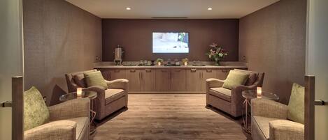 A modern, well-lit room with a wooden floor, two sofas facing a kitchen area with a TV screen, and a vase with flowers on the counter.