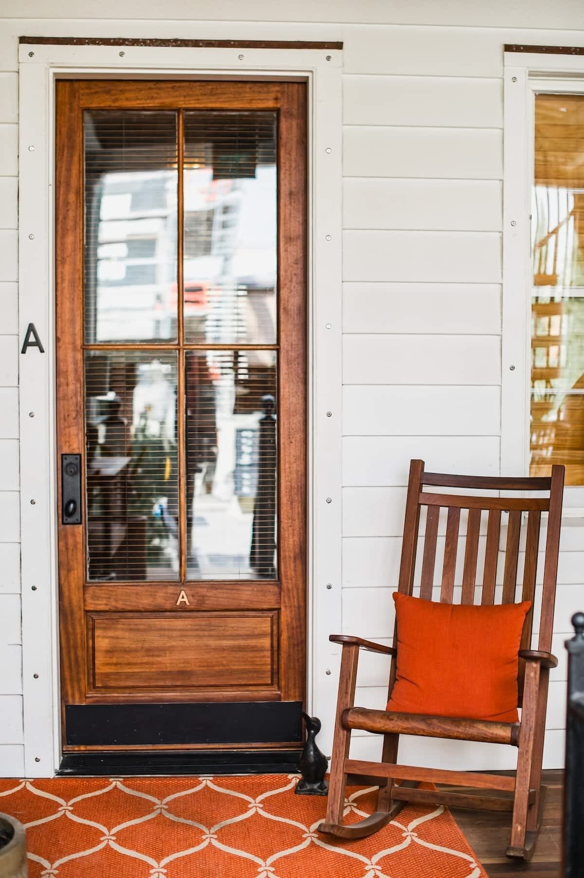 A wooden door with a glass panel, a wooden chair with an orange cushion, and a patterned rug on a wooden floor.