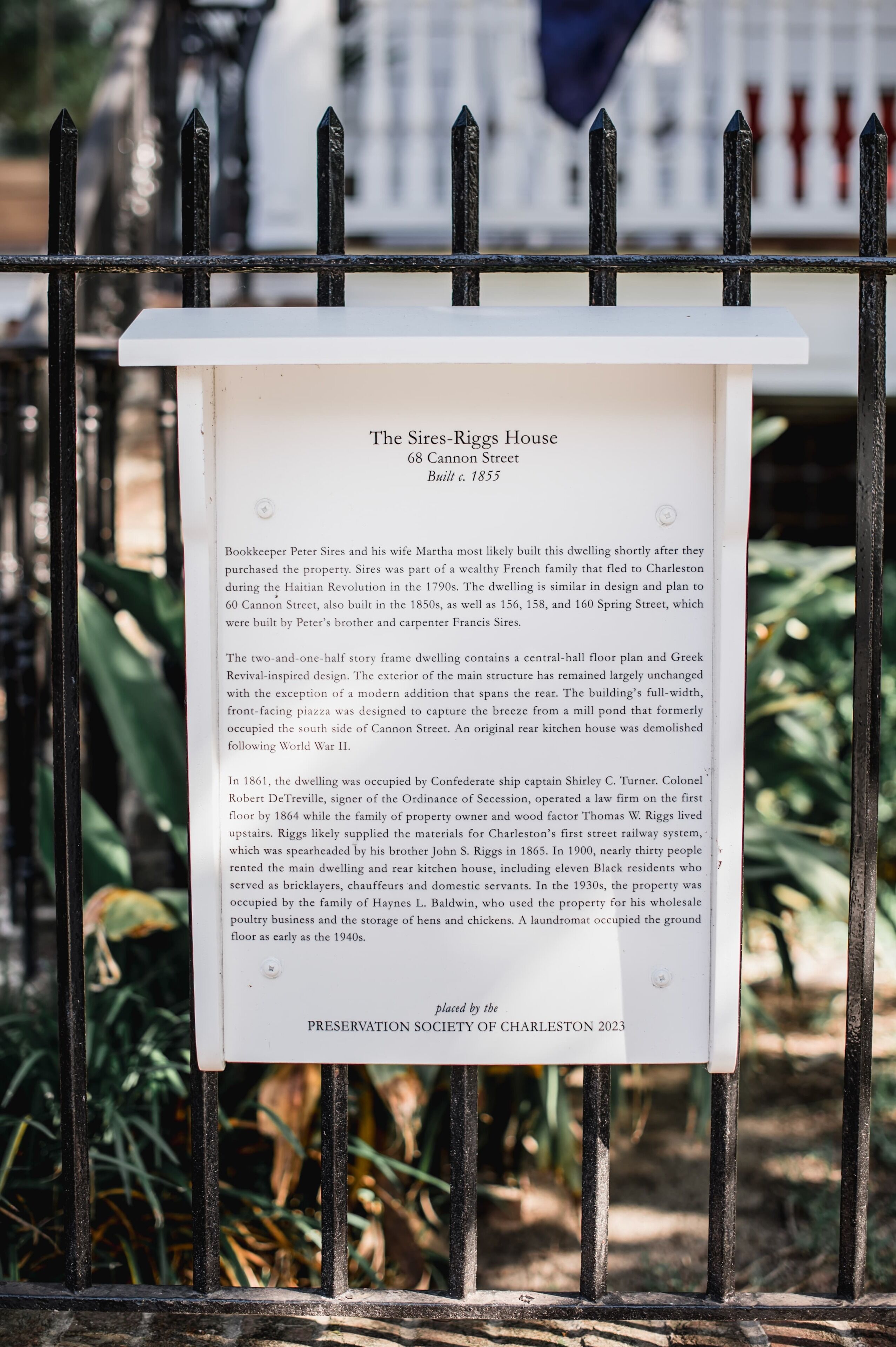 A historical plaque on a metal fence detailing The Sires-Riggs House, including its address, construction year, and historical significance.