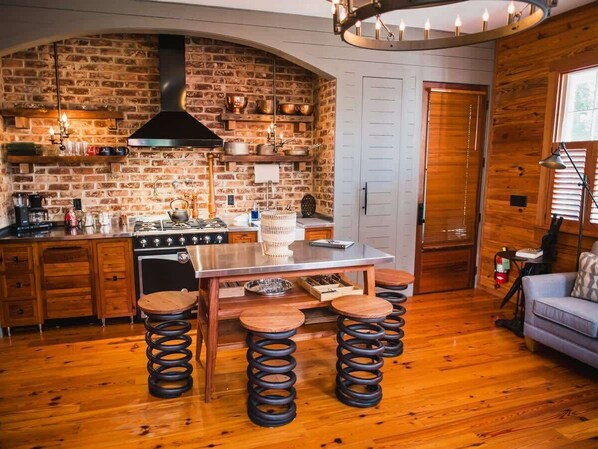 A rustic kitchen with a brick wall, wooden cabinets, a stainless steel island, and a hanging light fixture.