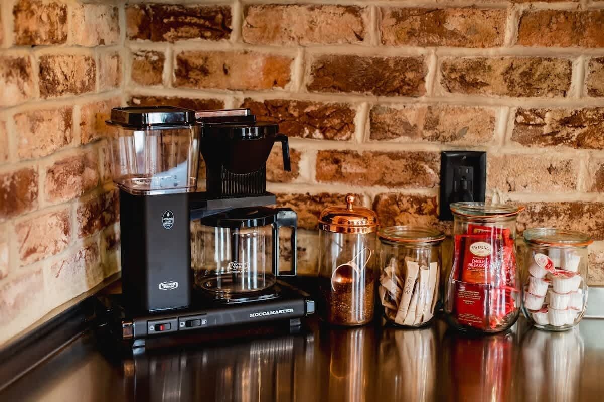 Une machine à café, une verseuse en verre et plusieurs bocaux en verre contenant des sachets de thé, ainsi qu’une boîte de thé English Breakfast de la marque Twinings, sont posés sur un comptoir adossé à un mur de briques.