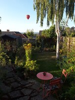 Un jardín con una mesa y sillas rojas, un globo aerostático al fondo y una variedad de plantas y árboles.