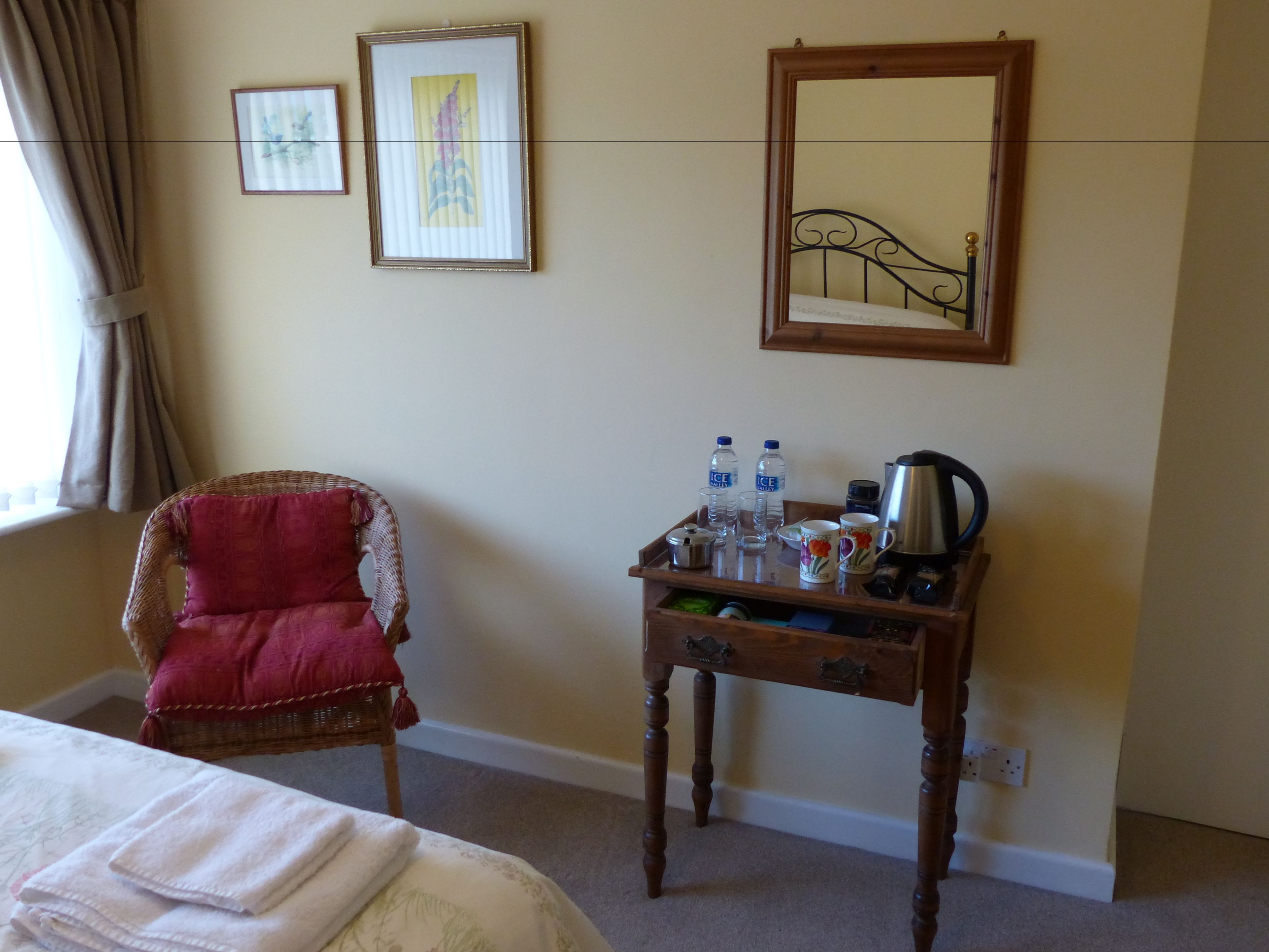 Una habitación de hotel con una cama, una silla, una mesita con botellas de agua, una cafetera y cuadros en la pared.