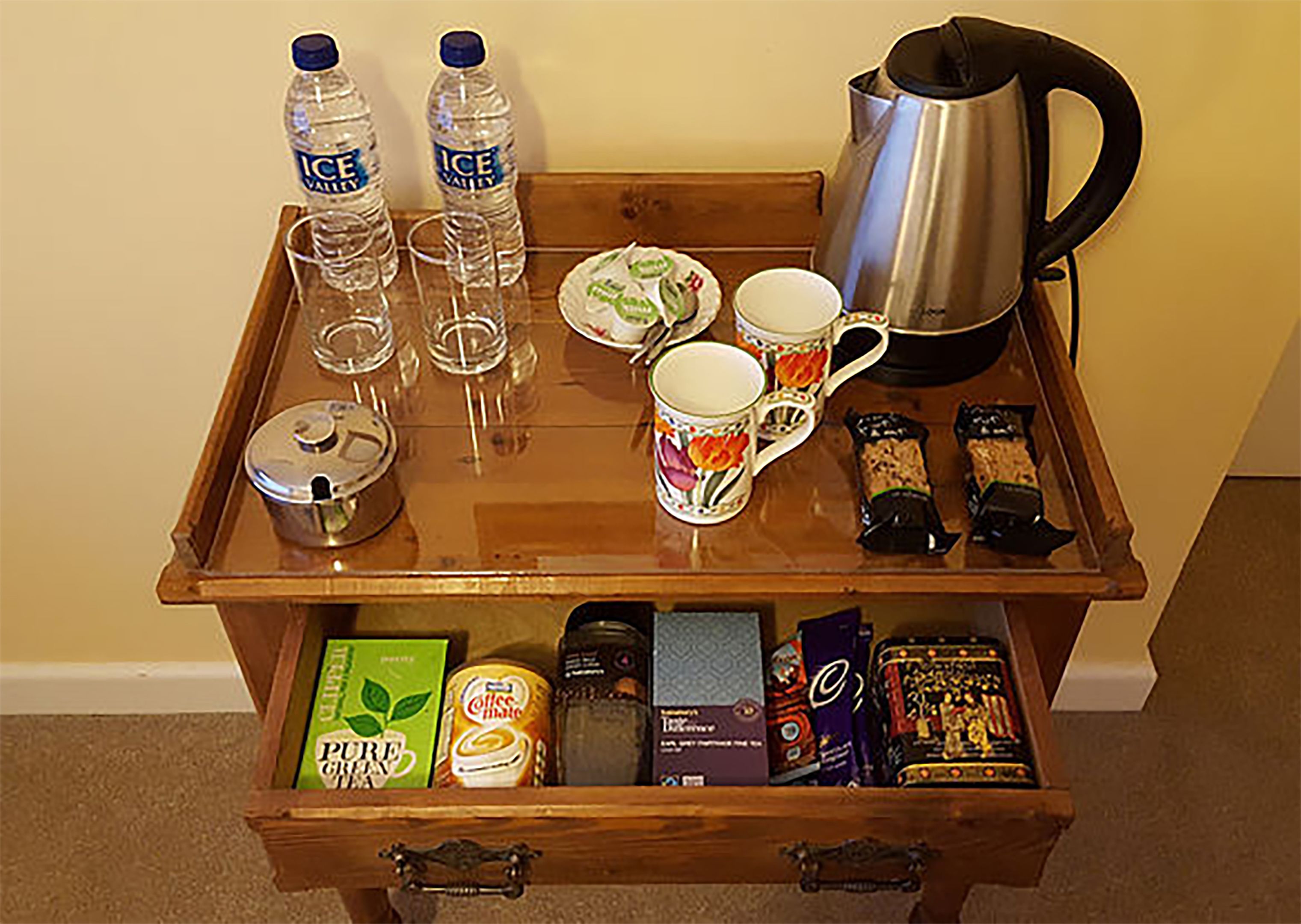 A wooden tray table with various items including two bottles of water, a coffee pot, a mug, and some snacks. Below the tray, there is a drawer with a green box and a can of coffee.