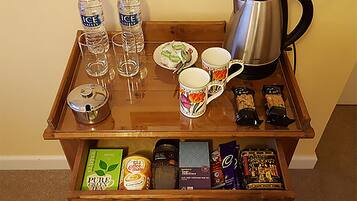 A wooden tray table with various items including two bottles of water, a coffee pot, a mug, and some snacks. Below the tray, there is a drawer with a green box and a can of coffee.