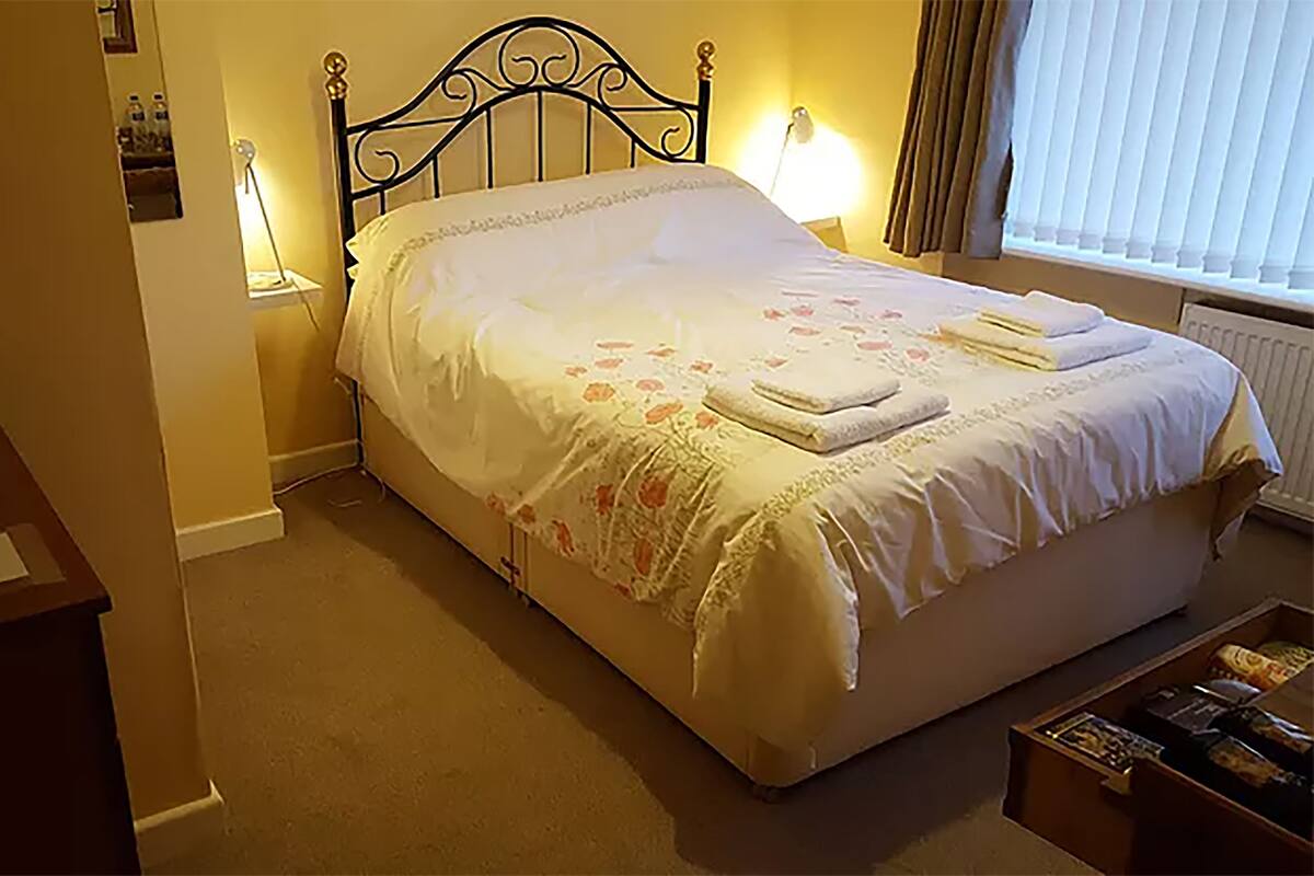 Een net opgemaakt bed met een bloemenprint sprei en twee kussens, een bedlamp en een klein tafeltje met een afstandsbediening.