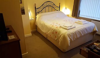 Una cama bien hecha con una colcha estampada con motivos florales y dos almohadas, una lámpara de noche y una mesita con un control remoto.