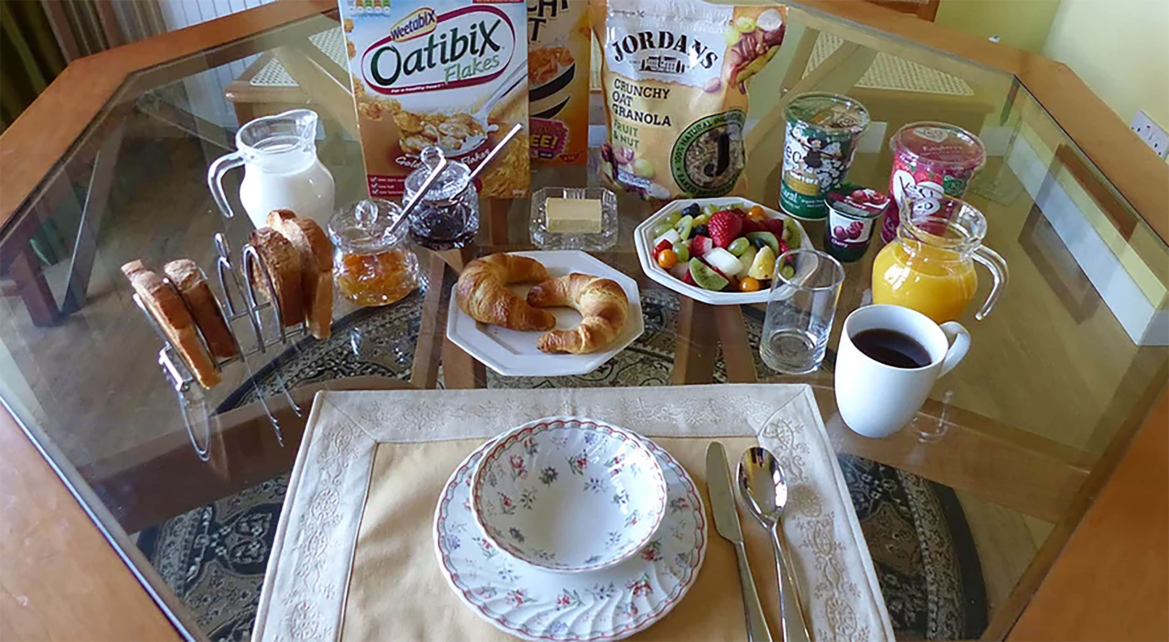 A breakfast table with a glass top, featuring a plate, croissant, jam, a box of Oatibix flakes, a bowl of fruit, a glass of orange juice, and a cup of coffee.