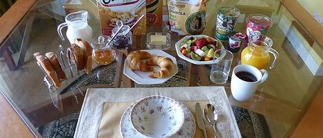 A breakfast table with a glass top, featuring a plate, croissant, jam, a box of Oatibix flakes, a bowl of fruit, a glass of orange juice, and a cup of coffee.